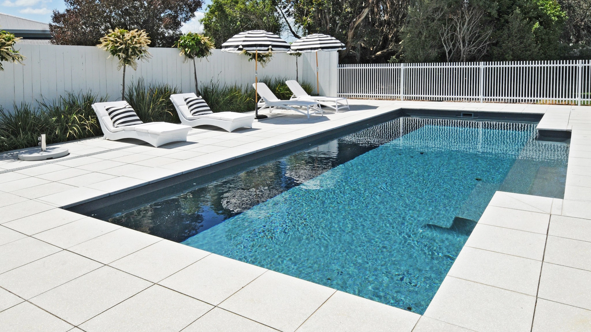 The Elegance 10.00m fibreglass swimming pool is installed in a metropolitan region of Martinborough, New Zealand. The house is surrounded by white tiles and natural greenery with mountains in the background. The pool colour is Graphite Grey, which stands out from the resort-like landscape and wooden house.