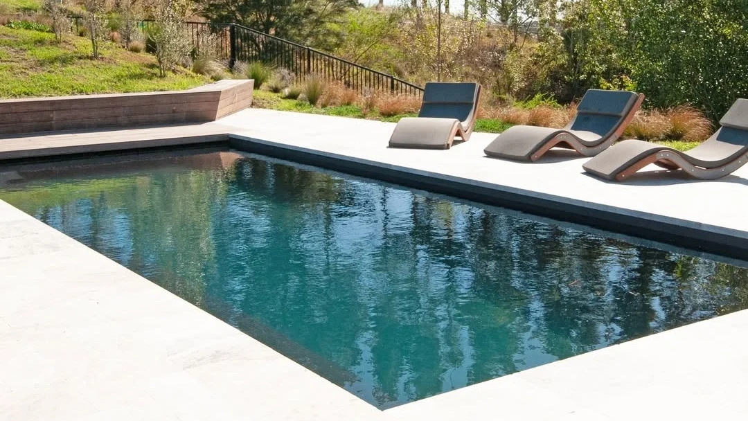 The Reflection with Auto Cover 9.90m fibreglass swimming pool is located in a residential backyard with contemporary pool accessories and landscaping surrounding it. It has an Ebony Blue colour that complements the greenery in the background and the pool chairs located next to it.