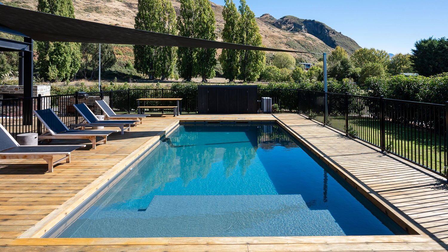 The Acclaim 10.20m fibreglass swimming pool is installed in a mountainous region of Central Otago, New Zealand. The house is surrounded by wooden tiles and natural greenery with mountains in the background. The pool colour is Graphite Grey, which stands out from the resort-like landscape and wooden house.