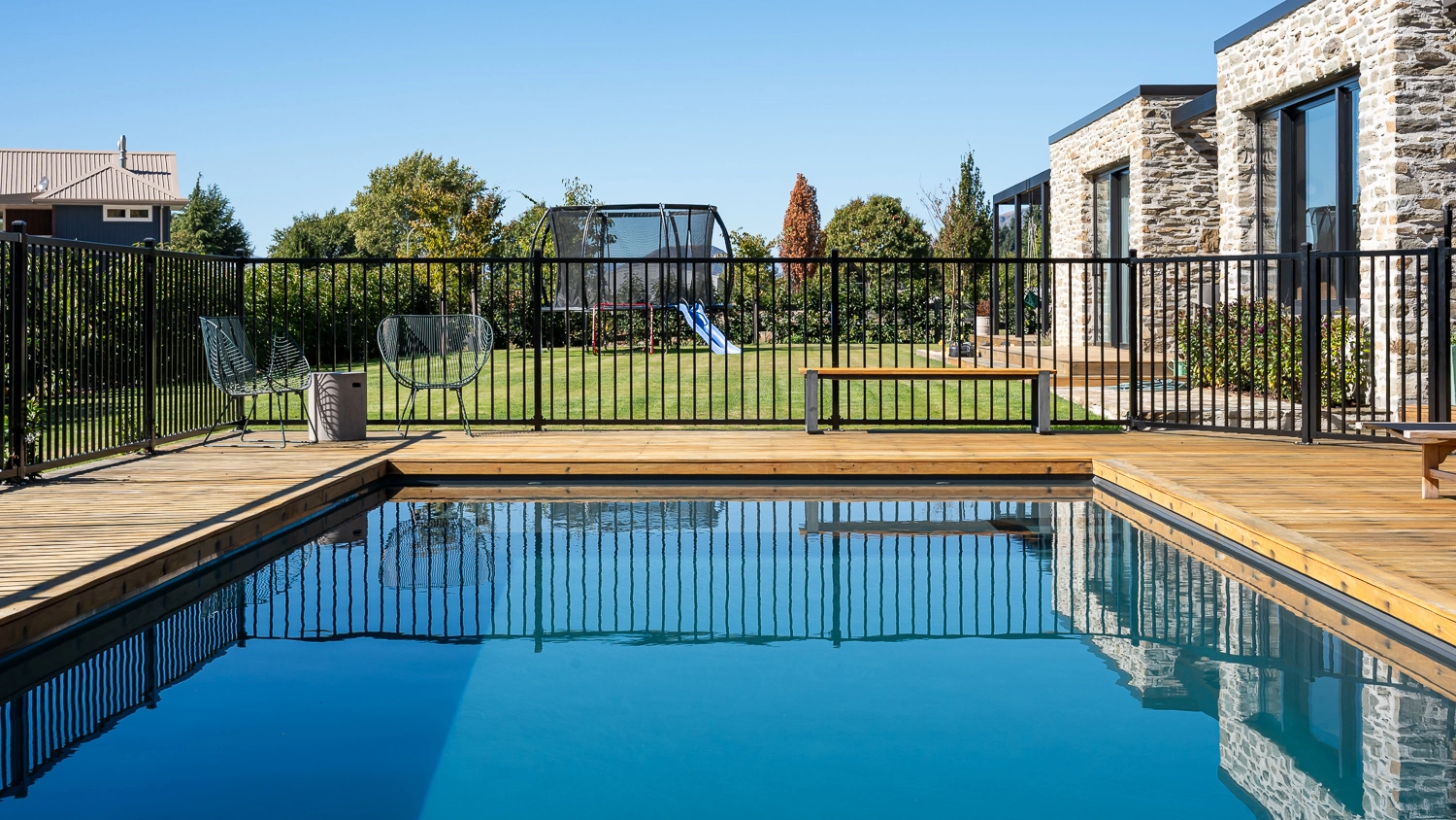 The Acclaim 10.20m fibreglass swimming pool is installed in a mountainous region of Central Otago, New Zealand. The house is surrounded by wooden tiles and natural greenery with mountains in the background. The pool colour is Graphite Grey, which stands out from the resort-like landscape and wooden house.