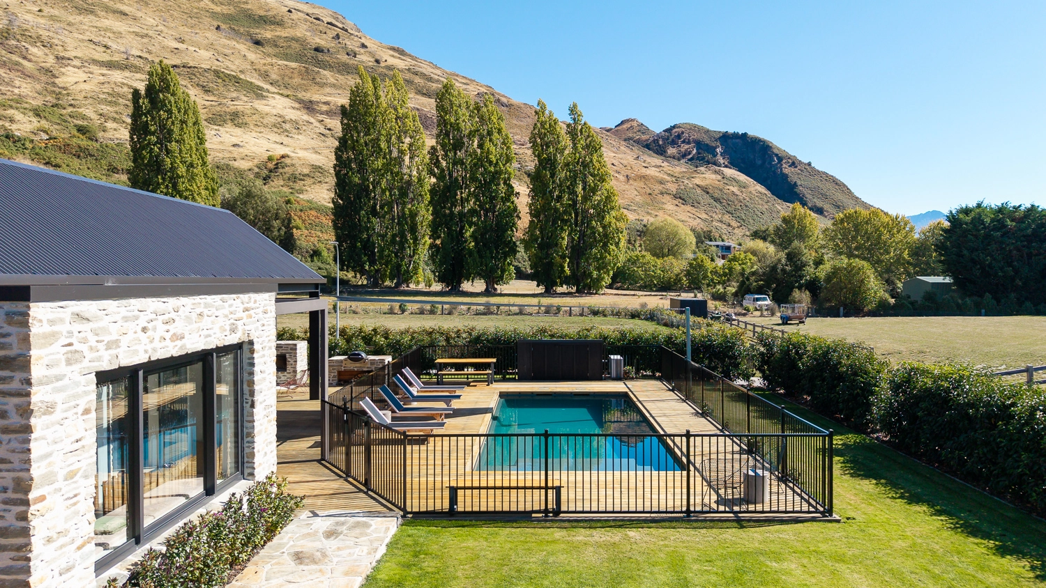 The Acclaim 10.20m fibreglass swimming pool is installed in a mountainous region of Central Otago, New Zealand. The house is surrounded by wooden tiles and natural greenery with mountains in the background. The pool colour is Graphite Grey, which stands out from the resort-like landscape and wooden house.