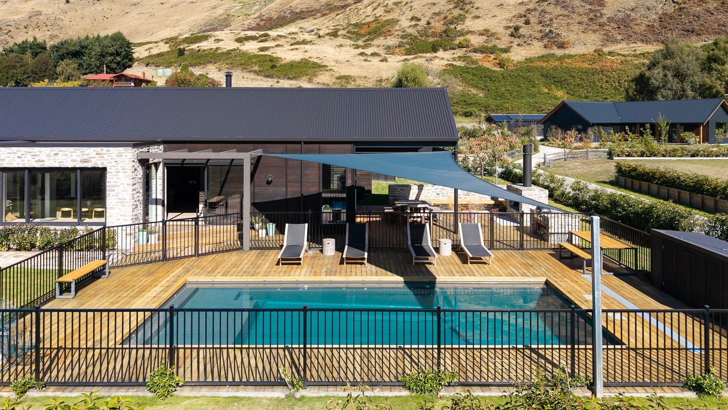 The Acclaim 10.20m fibreglass swimming pool is installed in a mountainous region of Central Otago, New Zealand. The house is surrounded by wooden tiles and natural greenery with mountains in the background. The pool colour is Graphite Grey, which stands out from the resort-like landscape and wooden house.