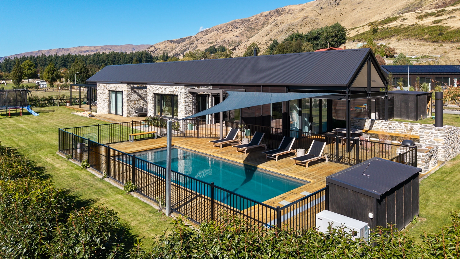 The Acclaim 10.20m fibreglass swimming pool is installed in a mountainous region of Central Otago, New Zealand. The house is surrounded by wooden tiles and natural greenery with mountains in the background. The pool colour is Graphite Grey, which stands out from the resort-like landscape and wooden house.