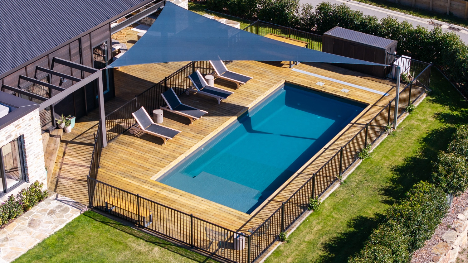 The Acclaim 10.20m fibreglass swimming pool is installed in a mountainous region of Central Otago, New Zealand. The house is surrounded by wooden tiles and natural greenery with mountains in the background. The pool colour is Graphite Grey, which stands out from the resort-like landscape and wooden house.