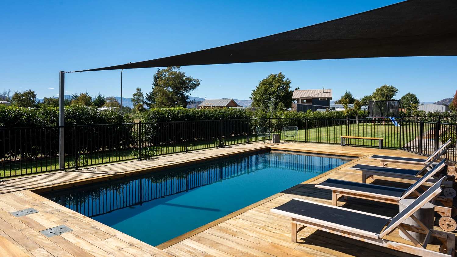 The Acclaim 10.20m fibreglass swimming pool is installed in a mountainous region of Central Otago, New Zealand. The house is surrounded by wooden tiles and natural greenery with mountains in the background. The pool colour is Graphite Grey, which stands out from the resort-like landscape and wooden house.