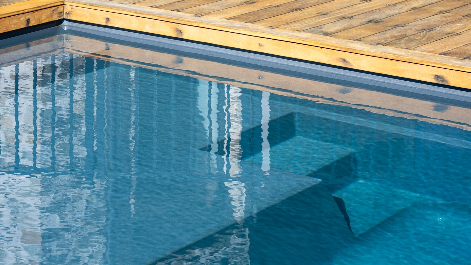 The Acclaim 10.20m fibreglass swimming pool is installed in a mountainous region of Central Otago, New Zealand. The house is surrounded by wooden tiles and natural greenery with mountains in the background. The pool colour is Graphite Grey, which stands out from the resort-like landscape and wooden house.