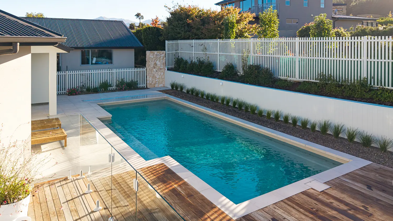 The Elegance 10.00m fibreglass swimming pool is installed in a metropolitan region of Central Otago, New Zealand. The house is surrounded by wooden tiles and natural greenery with mountains in the background. The pool colour is Diamond Sand, which stands out from the resort-like landscape and wooden house.
