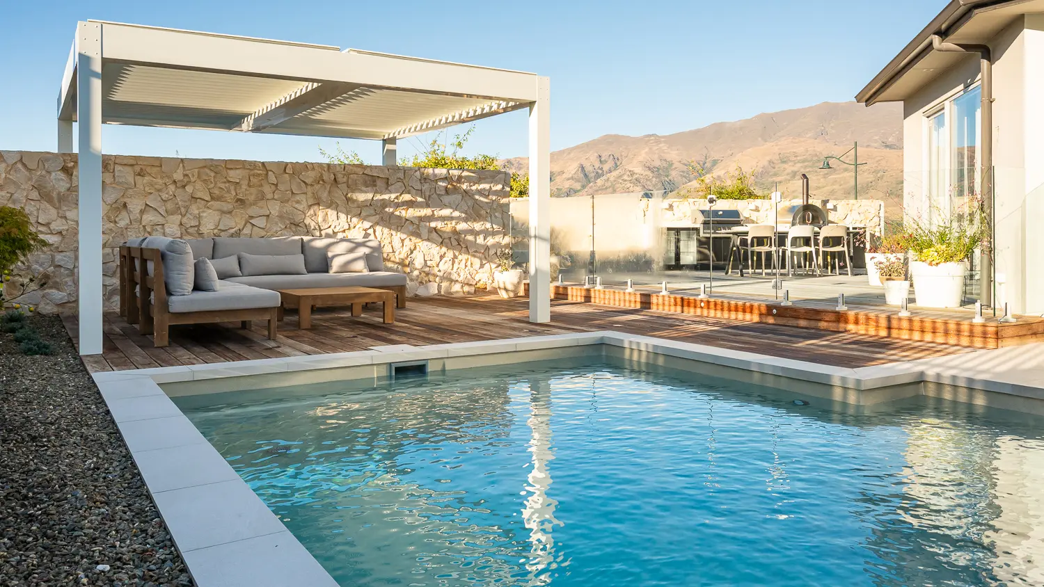 The Elegance 10.00m fibreglass swimming pool is installed in a metropolitan region of Central Otago, New Zealand. The house is surrounded by wooden tiles and natural greenery with mountains in the background. The pool colour is Diamond Sand, which stands out from the resort-like landscape and wooden house.