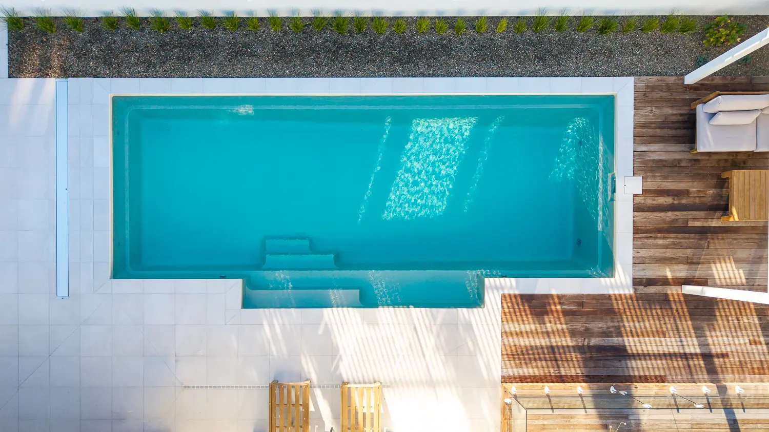 The Elegance 10.00m fibreglass swimming pool is installed in a metropolitan region of Central Otago, New Zealand. The house is surrounded by wooden tiles and natural greenery with mountains in the background. The pool colour is Diamond Sand, which stands out from the resort-like landscape and wooden house.