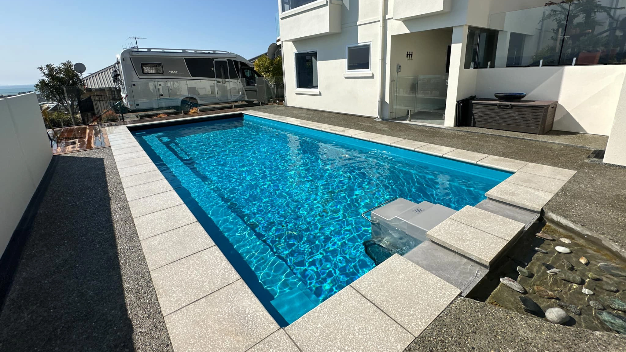 The Reflection with Auto Cover 9.90m fibreglass swimming pool is installed in a metropolitan region of Stoke, New Zealand. The house is surrounded by grey tiles, a small waterfall and natural greenery with mountains in the background. The pool colour is Crystal Blue, which stands out from the tranquil landscape and white house.