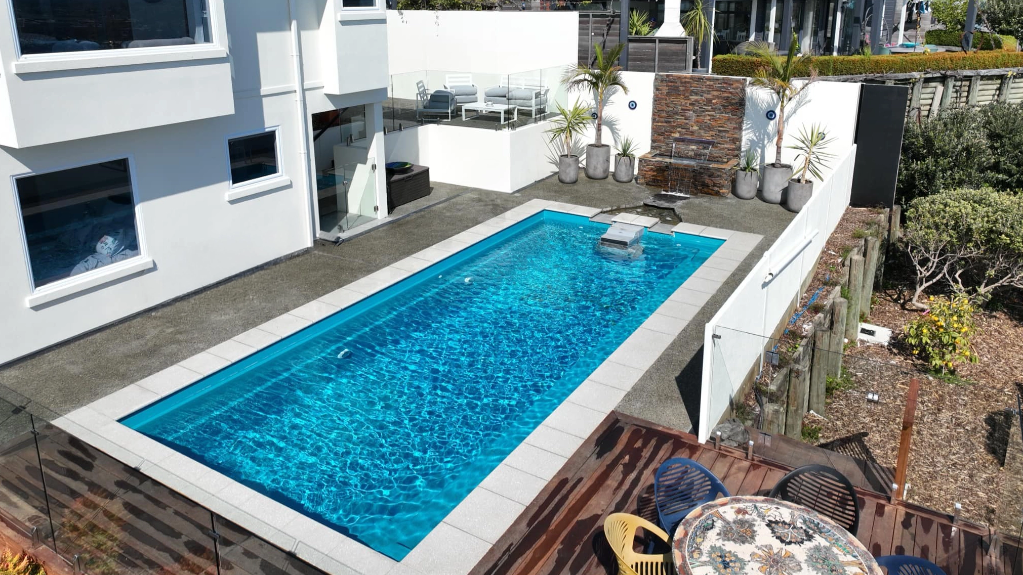 The Reflection with Auto Cover 9.90m fibreglass swimming pool is installed in a metropolitan region of Stoke, New Zealand. The house is surrounded by grey tiles, a small waterfall and natural greenery with mountains in the background. The pool colour is Crystal Blue, which stands out from the tranquil landscape and white house.