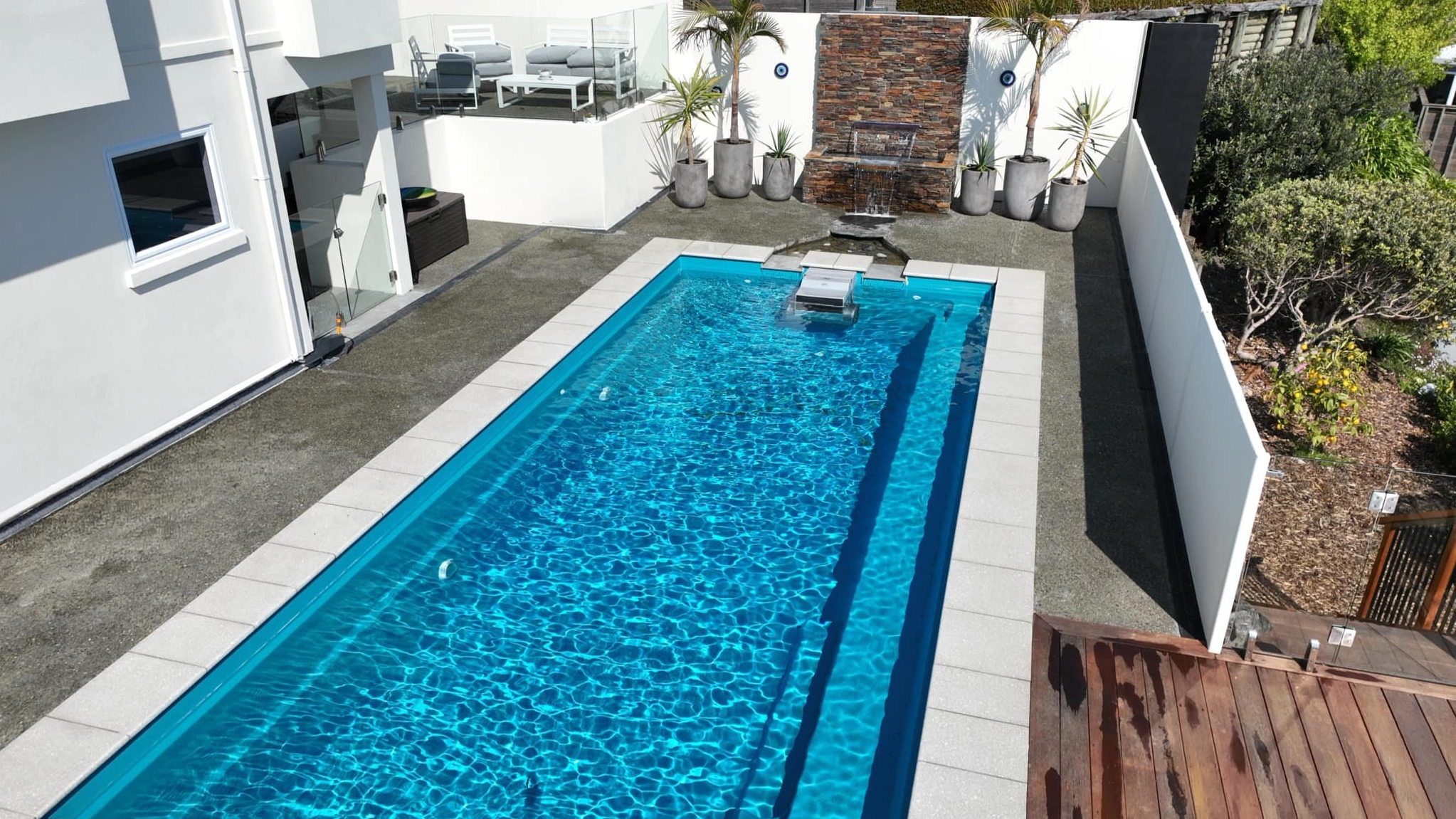 The Reflection with Auto Cover 9.90m fibreglass swimming pool is installed in a metropolitan region of Stoke, New Zealand. The house is surrounded by grey tiles, a small waterfall and natural greenery with mountains in the background. The pool colour is Crystal Blue, which stands out from the tranquil landscape and white house.