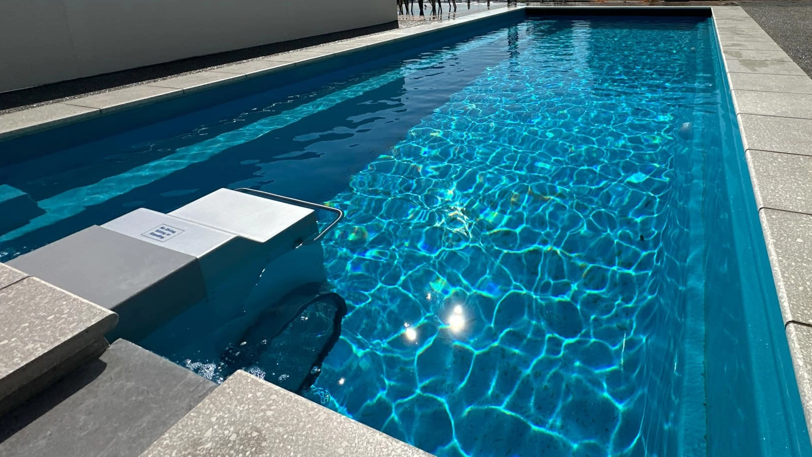 The Reflection with Auto Cover 9.90m fibreglass swimming pool is installed in a metropolitan region of Stoke, New Zealand. The house is surrounded by grey tiles, a small waterfall and natural greenery with mountains in the background. The pool colour is Crystal Blue, which stands out from the tranquil landscape and white house.